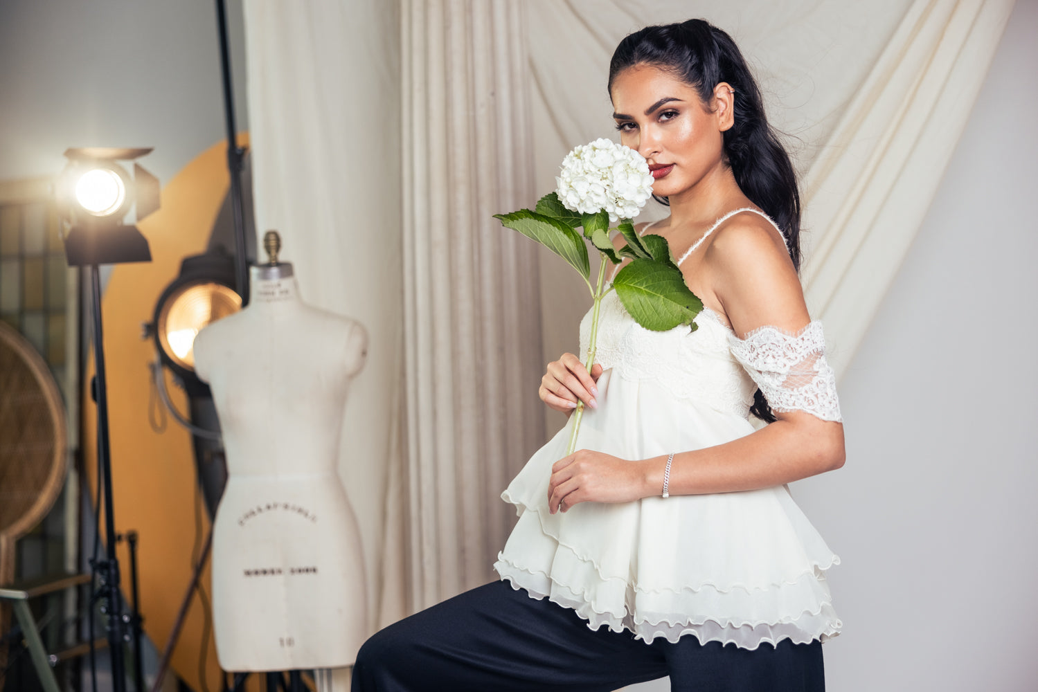 Woman in a white top holding flowers in a studio setting with a mannequin and lighting equipment.