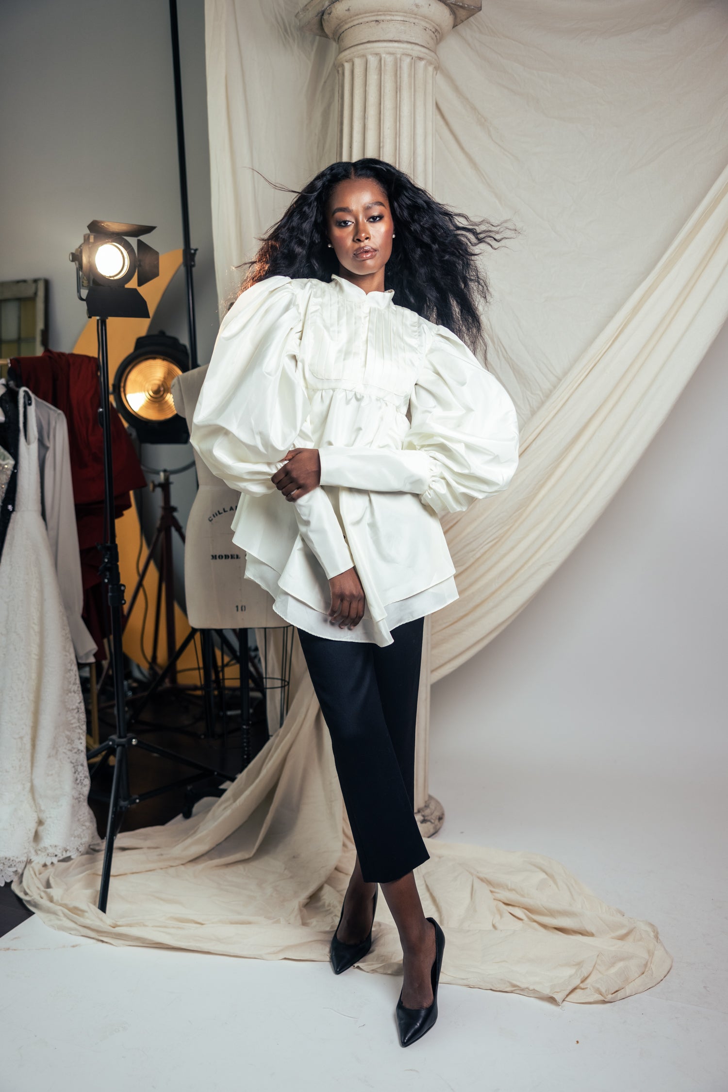 Woman in a white blouse and black skirt standing in a studio setting with draped fabric and lighting equipment.