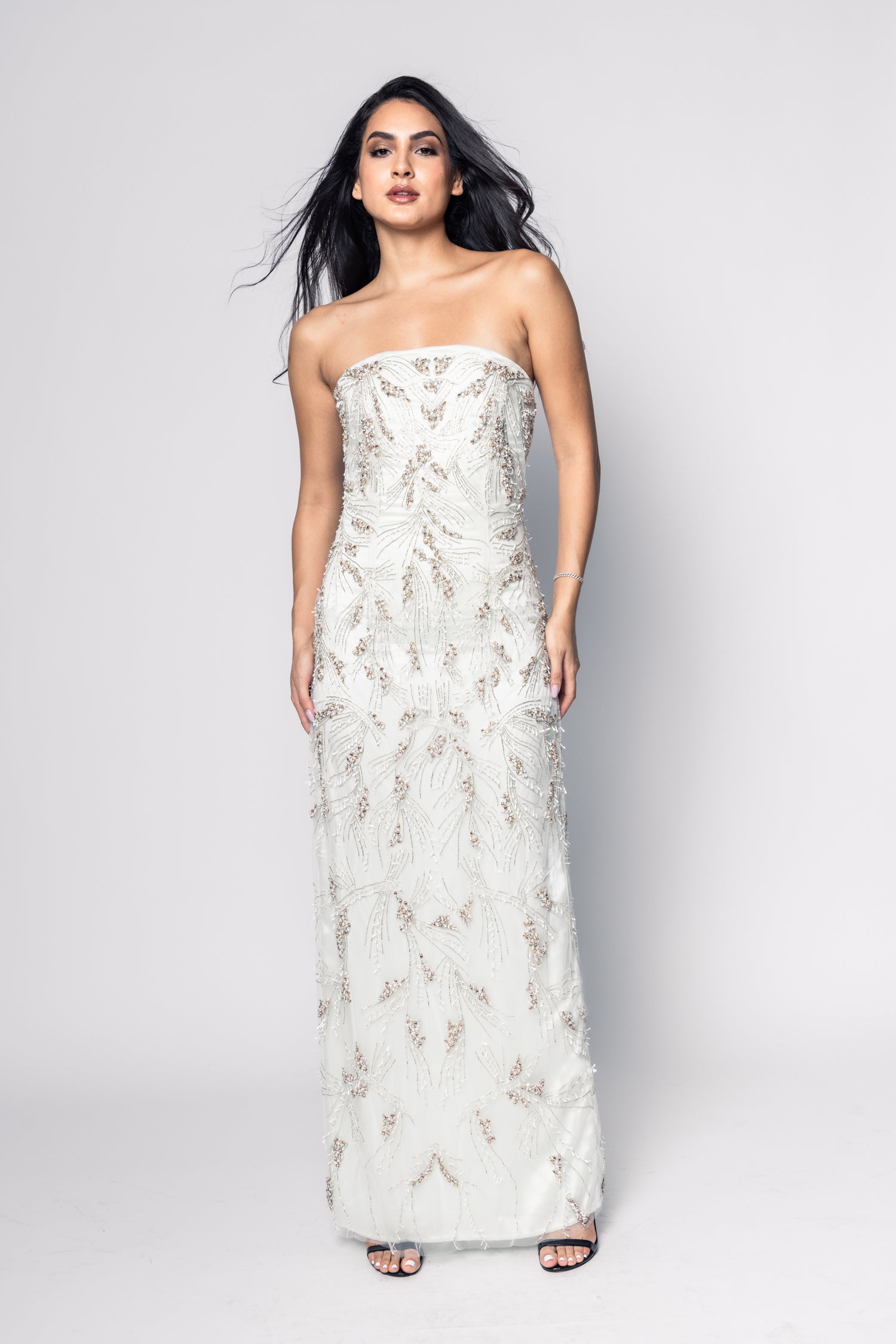 Woman wearing a strapless, long, white embroidered dress against a plain background