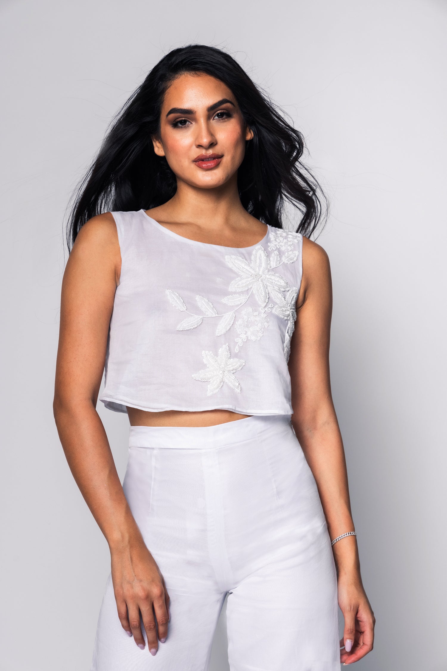 Woman wearing a white sleeveless top with floral details and white pants against a gray background