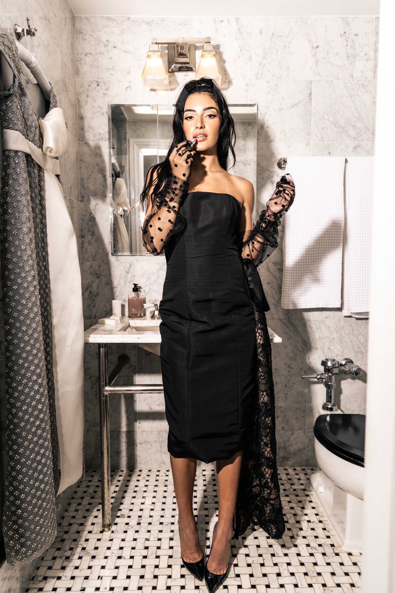 Gorgeous model in chic black embellished Sujata cocktail dress with elbow-length tulle gloves and a velvet bow in her hair putting on lipstick in a bathroom