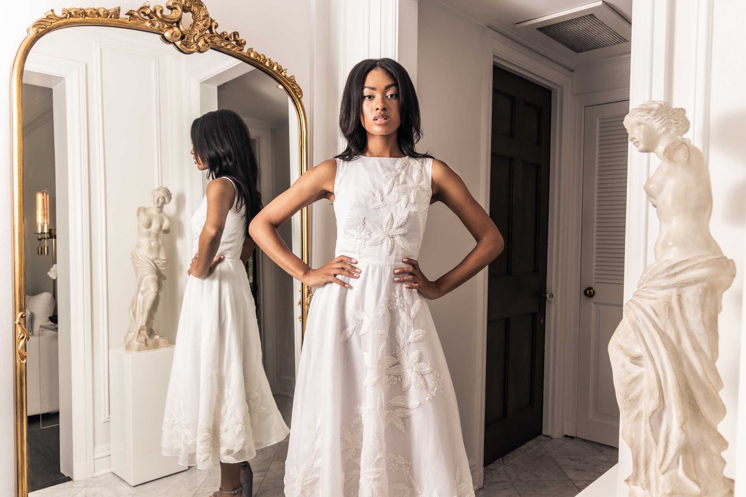 Beautiful model in an embroidered, beaded, ornate Sujata Gazder tea-length dress in front of an ornate mirror and statue