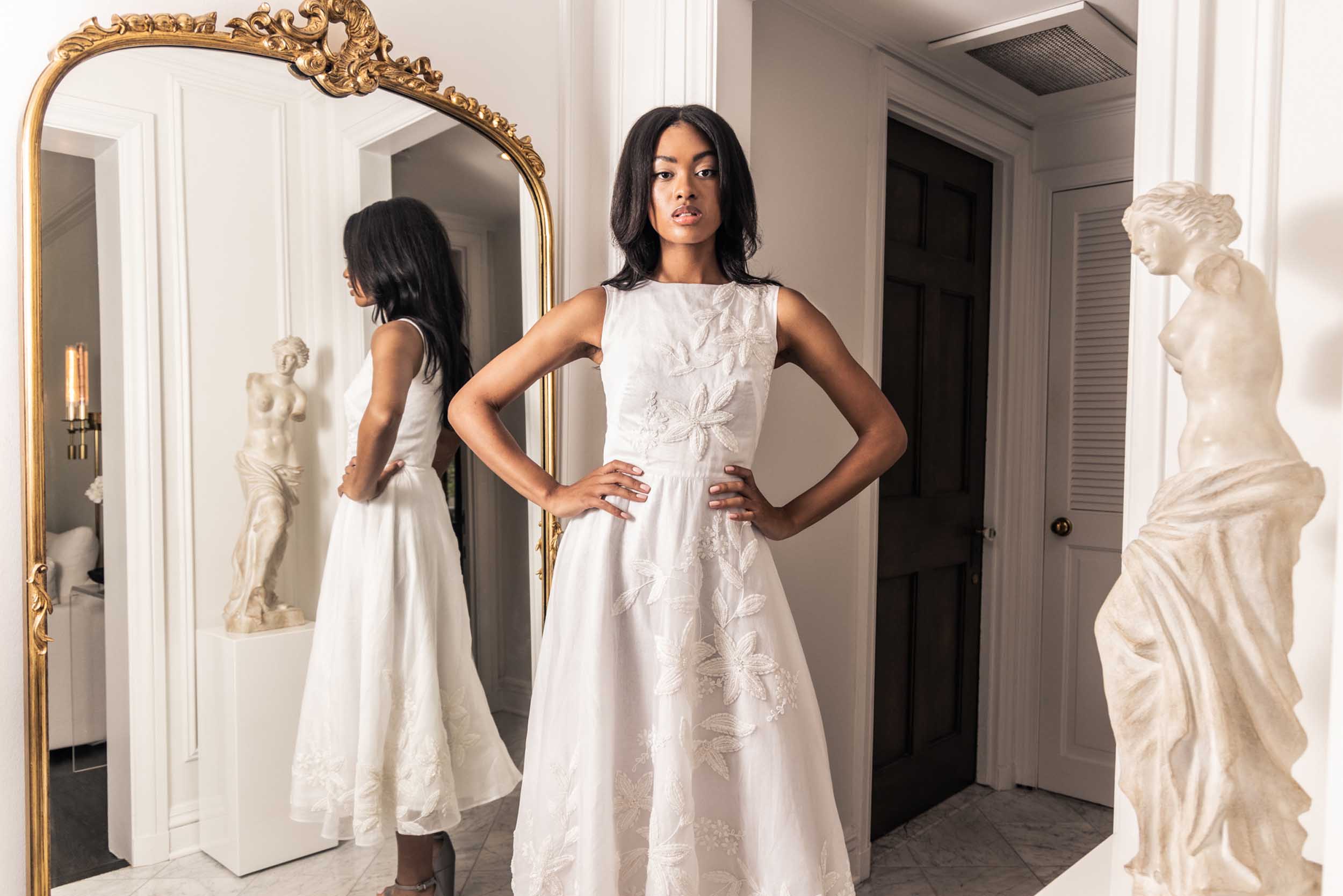 Gorgeous model in a beaded white tea-length Sujata Gazder gown in a chic setting with ornate mirror and statue