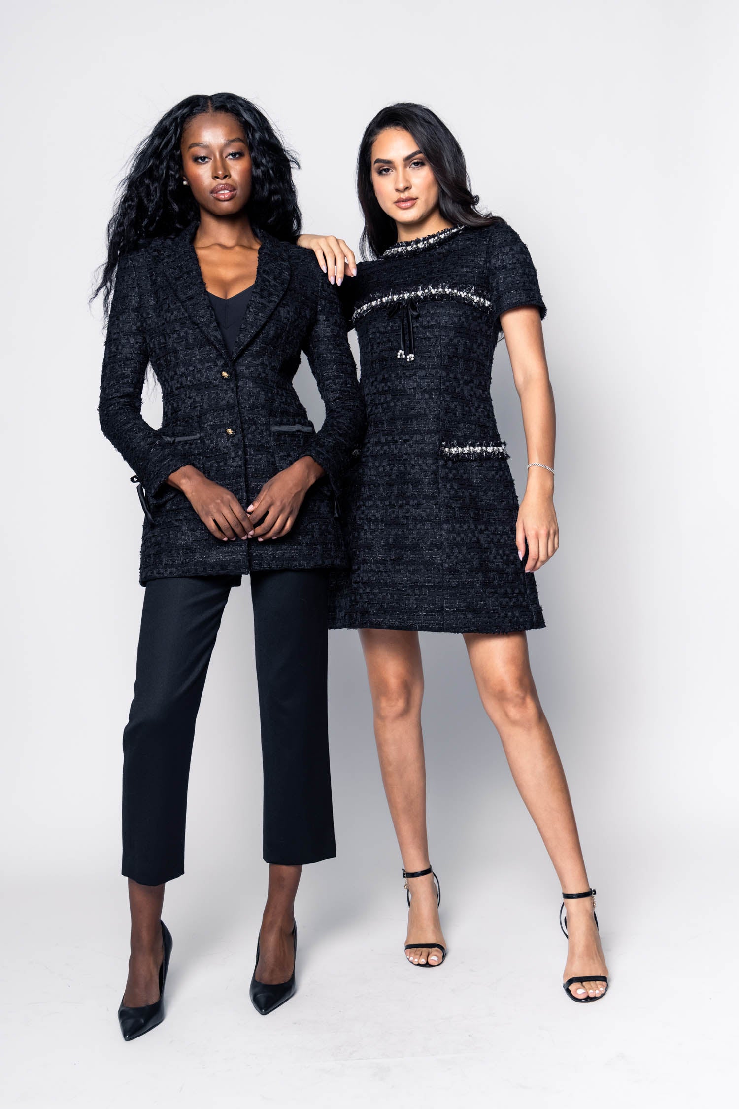 Two women wearing black tweed suit and a boucle short black dress on a white background