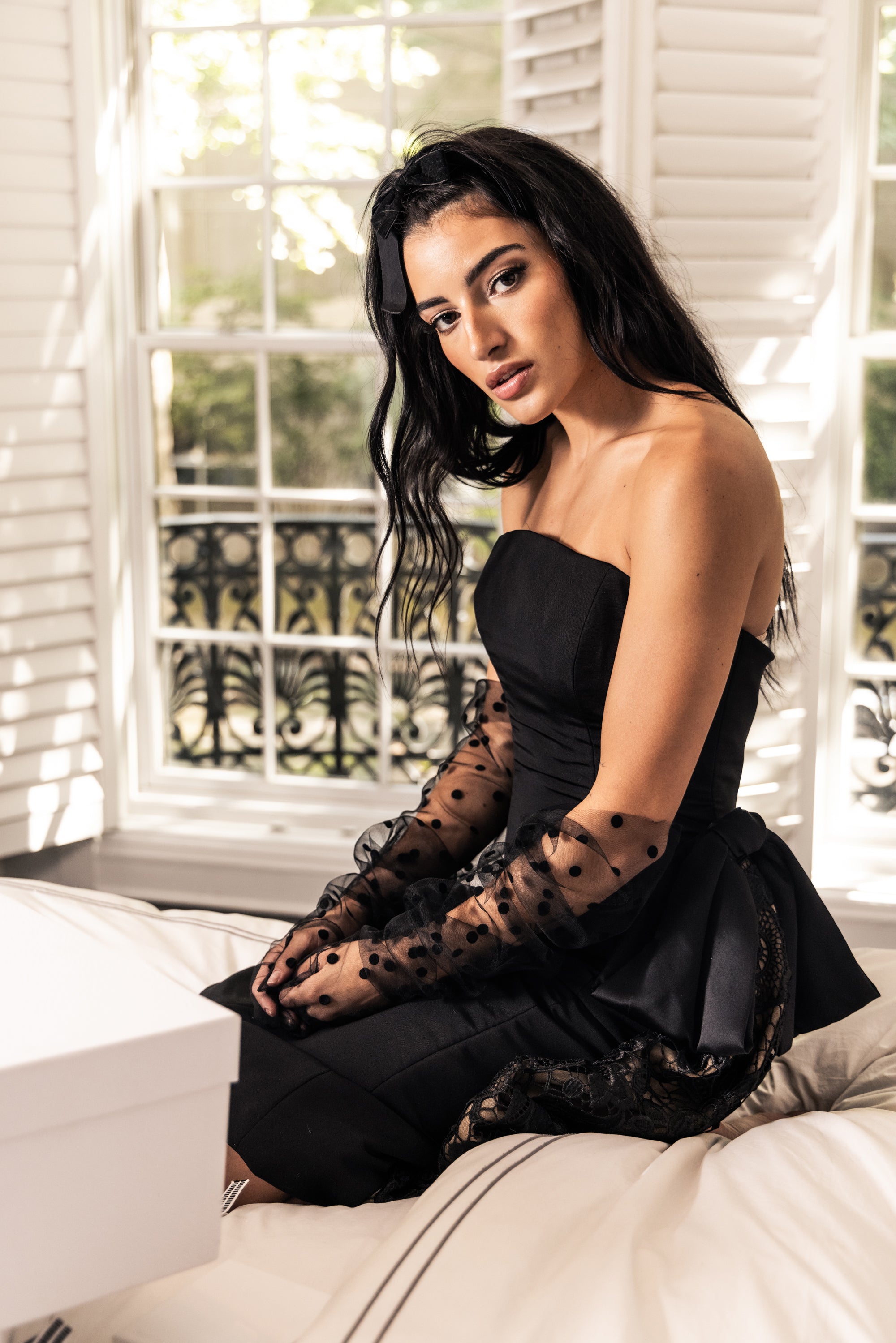 Gorgeous model in chic black embellished Sujata cocktail dress with elbow-length tulle gloves and a velvet bow in her hair kneeling on a bed with packages
